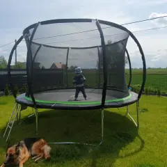 Dziecko w czapce na trampolinie, pies odpoczywa na trawie. Czas na wesołe zabawy na trampolinie w ogrodzie.