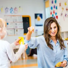 Nauczycielka z uśmiechem prowadzi zajęcia, budując poczucie bezpieczeństwa u dziecka. W tle kolorowe liczby i dekoracje.