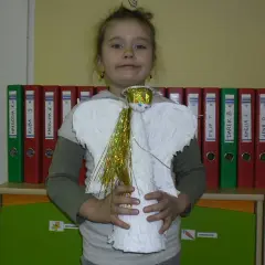 Dziecko trzyma anioła, pracę plastyczną przestrzenną. Skrzydła i tułów wykonane z białej bibuły, główka ozdobiona złotą aureolą i błyszczącymi włosami.