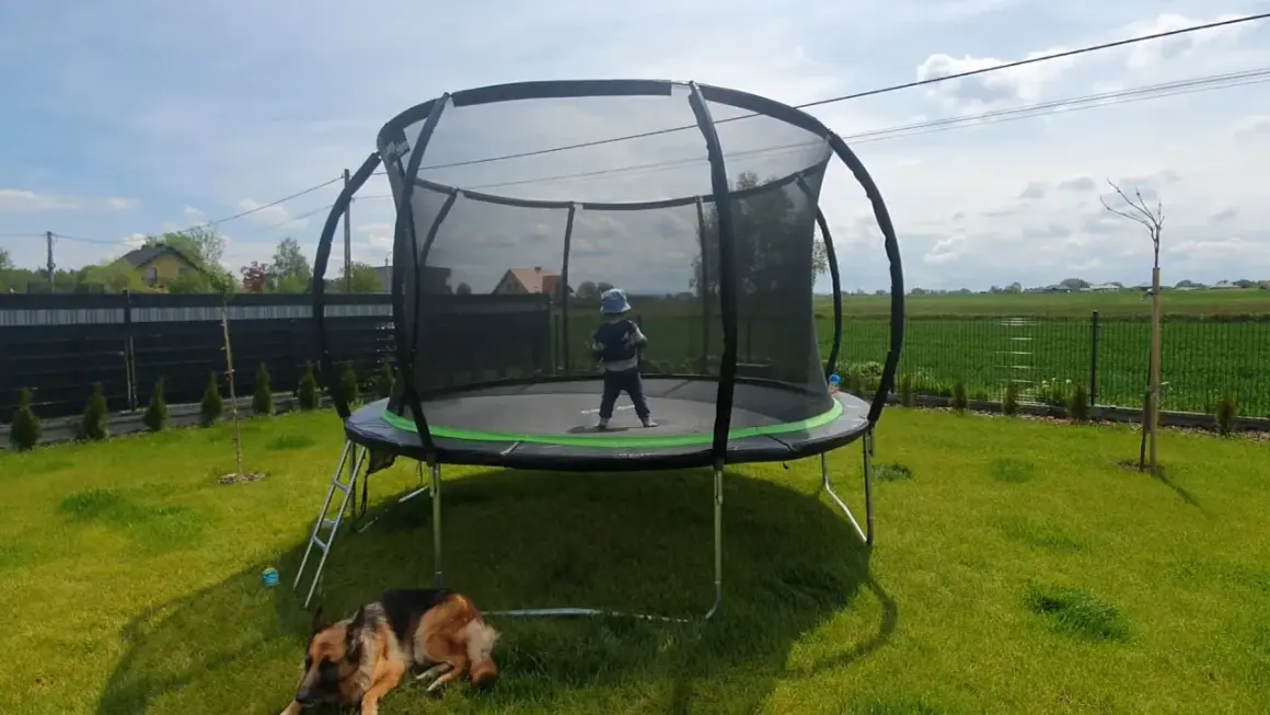 Dziecko w czapce na trampolinie, pies odpoczywa na trawie. Czas na wesołe zabawy na trampolinie w ogrodzie.