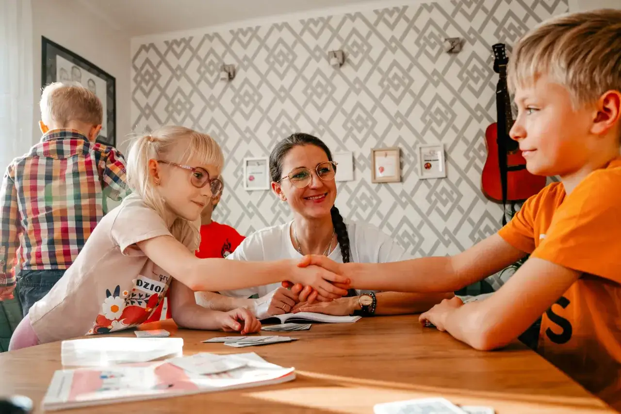 Dzieci uczą się radzić sobie z emocjami. 6 latek ściska dłoń chłopca, a kobieta uśmiecha się.