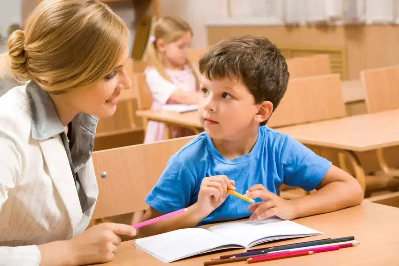 Nauczycielka wspiera chłopca z specjalnymi potrzebami edukacyjnymi w nauce. W tle widać inną uczennicę.